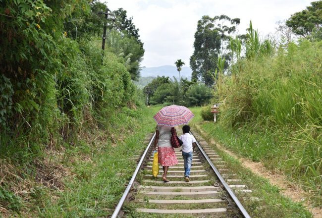 Two Sri Lankan people walking along a train track with their backs to us, one holding a sun parasol / umbrella